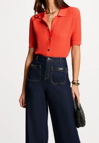 Czerwona dzianinowa bluzka o fakturze z krótkimi rękawami i guzikami, zestawiona z wysokim stanem szerokich spodni z ciemnego dżinsu z ozdobnymi elementami metalowymi.