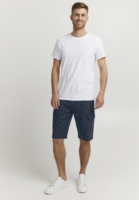 Un t-shirt blanc à col rond associé à un short en cargo bleu marine avec plusieurs poches. Chaussures : baskets blanches. Design simple et décontracté.