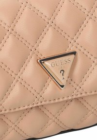 Beige, gesteppte Lederoberfläche mit einem dreieckigen Metallabzeichen, das "GUESS U.S.A" und ein Fragezeichen-Logo eingraviert hat.