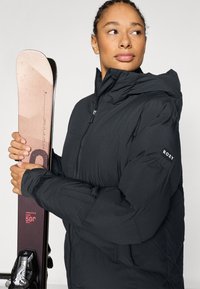 Schwarze isolierte Jacke mit Kapuze und Roxy-Logo am Ärmel; hält pinke und braune Skier mit strukturierter Oberfläche.