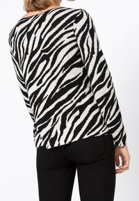 Svartvit zebra-randig tröja med långa ärmar och bred halsringning, visad bakifrån, tillsammans med svarta byxor.