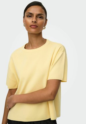 Femme aux cheveux tirés en arrière portant un haut à manches courtes jaune pâle et des boucles d'oreilles en créole dorées, posant avec un bras croisé sur la taille.