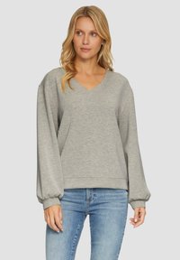 Grijze pullover sweatshirt met een V-hals, lange volumineuze mouwen en geribde manchetten. Lichtgewicht stof met een zachte textuur. Ggedragen met blauwe jeans.