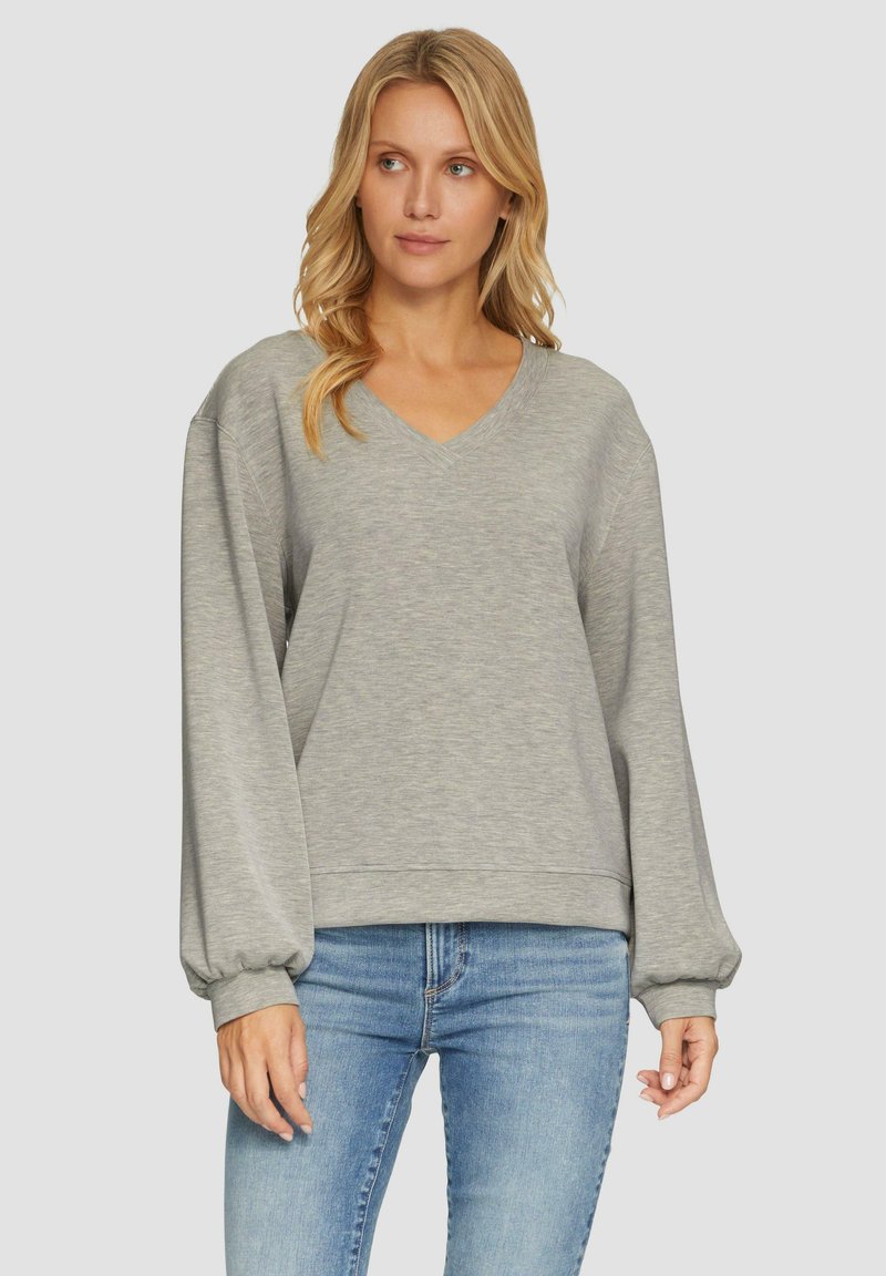 Grijze pullover sweatshirt met een V-hals, lange volumineuze mouwen en geribde manchetten. Lichtgewicht stof met een zachte textuur. Ggedragen met blauwe jeans.