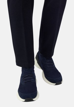 Marineblaue Strick-Sneaker mit dicken weißen Sohlen, getragen mit schwarzen Hosen, fotografiert von vorne auf Knöchelhöhe.