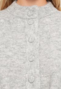 Γκρι πλέκτο ζακέτα με ψηλό γιακά και τρία στρογγυλά κουμπιά στο μπροστινό μέρος. Μαλακή υφή με ελαφρώς χνουδωτό φινίρισμα.