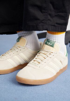 Personne portant des baskets Adidas Gazelle crème avec des semelles en gomme et des chaussettes blanches à liseré jaune, debout sur un sol bleu clair.