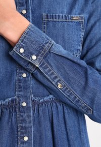 Denim-paita, jossa on sopiva muotoilu ja kerätty vyötärö. Vaaleansininen väri, hopeiset nepparit ja rintatasku, jossa on logotägi.