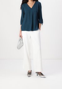 Błękitna bluzka z dekoltem w serek i rękawami 3/4, zestawiona z białymi spodniami o szerokich nogawkach i srebrnymi metalicznymi butami, trzymając srebrną torebkę.