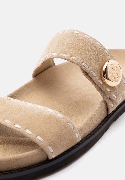 Μπεζ σουέντ σανδάλι με δύο ραμμένους ιμάντες, ένα χρυσό διακοσμητικό κουμπί και μαύρη σόλα, εμφανιζόμενο σε κοντινή λεπτομέρεια.
