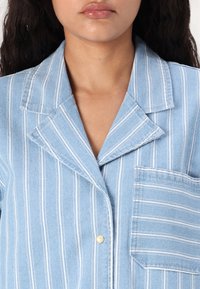 Camicia a righe blu chiaro con colletto, taschino sul petto e chiusura a bottoni. Tessuto testurizzato con dettagli di cucitura visibili.