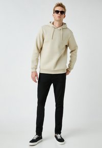 Sudadera beige con cordones, jeans negros ajustados y zapatillas negras con suelas blancas, llevadas con gafas de sol oscuras.