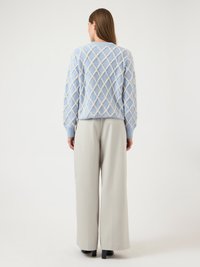 Γυναίκα με γυρισμένη την πλάτη, φορώντας ανοιχτό μπλε πουλόβερ με λευκό διαμαντένιο σχέδιο και μπεζ παντελόνια φαρδιάς γραμμής με μαύρα παπούτσια, σε λευκό φόντο.