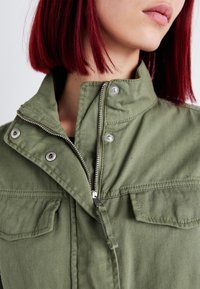 Oliivinvihreä takki, jossa on korkea kaulus, hopeiset painonapit ja etuvetoketju. Siinä on kaksi rintataskua ja teksturoitu kangas.