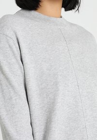 Kobieta nosząca jasnoszary sweter typu crewneck z centralnym pionowym szwem na przodzie.