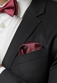 Musta puku, jossa on huivi-kaulus, valkoinen paita ja burgundinpunainen satiininen solmio. Rintataskuliina esittelee paisley-kuviota syvän punaisena ja sinisenä.