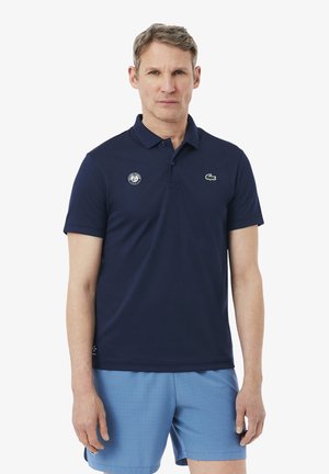 Uomo con polo blu navy con loghi e pantaloncini sportivi azzurri, in piedi contro uno sfondo bianco semplice.
