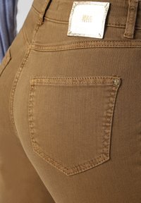 Jeans en denim marron mettant en avant une seule poche arrière avec des coutures contrastées et une étiquette de marque. Le tissu semble lisse et texturé.