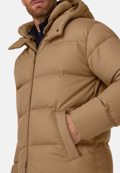 Homme portant une doudoune beige avec une fermeture éclair sur le devant et la main dans la poche latérale, visage partiellement visible et chemise bleu marine en dessous.