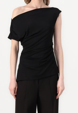 Vrouw draagt een zwarte asymmetrische off-shoulder top met korte mouwen, gecombineerd met een zwarte broek, staand tegen een effen achtergrond.