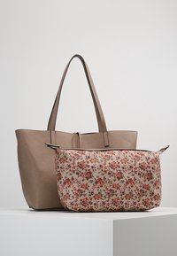 Ruskea nahkainen olkalaukku, jossa on kaksi kahvaa, yhdistettynä beigeen kukkakuosisella kankaiselle laukulle, jossa on punaisia ja vihreitä yksityiskohtia.