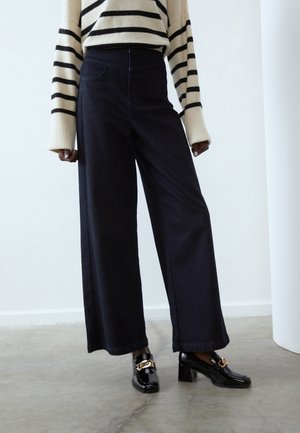 Osoba ubrana w czarne spodnie o szerokich nogawkach, czarne mokasyny na obcasie z ozdobnym złotym łańcuchem oraz beżowy sweter z czarnymi poziomymi paskami.