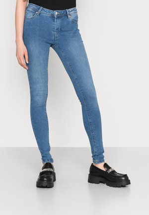 Frau trägt hellblaue Skinny-Jeans und schwarze Plateau-Loafer mit Silberketten-Detail, steht vor einem schlichten hellen Hintergrund.