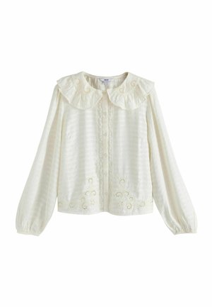 Vrouw zittend met gekruiste benen op een beige stoel, draagt een witte blouse met kanten details en olijfgroene wijde broek met bruine schoenen.