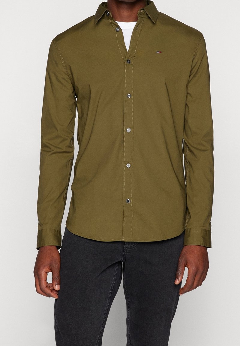 Homme portant une chemise olive à manches longues boutonnée sur un t-shirt blanc et un pantalon noir, debout, bras détendus.
