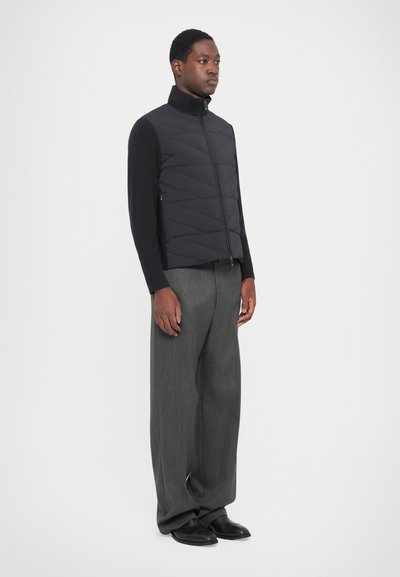 Black quilted vest with high collar, zippers, and a fitted design, paired with loose gray trousers and black shoes.