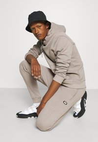 Light beige hoodie and matching sweatpants with ribbed cuffs. Black bucket hat and sporty sneakers with white and black accents. Close-up pose.