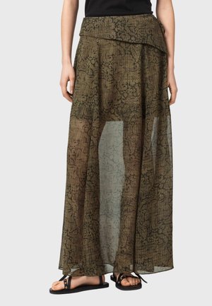 Femme portant une jupe longue en voile vert olive à motifs floraux noirs et des sandales plates noires, debout devant un fond uni.