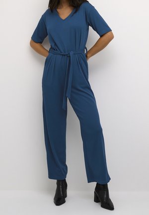 Blue ribbed jumpsuit with short sleeves, V-neck, belted waist, and wide pants; worn with black heeled ankle boots.