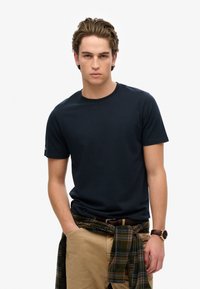 Marineblå crew-neck T-shirt i bomuld med korte ærmer, parret med khakifarvede bukser og en grønternet flannelbundet om taljen. Brun urskov synlig.