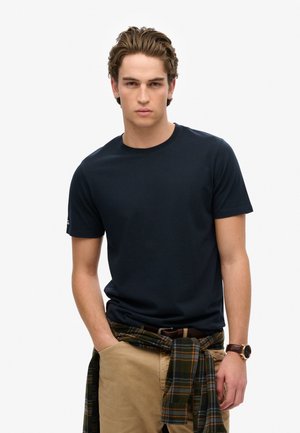 Navy crew-neck T-shirt in cotton with short sleeves, paired with khaki pants and a green plaid flannel tied around the waist. Brown watch visible.