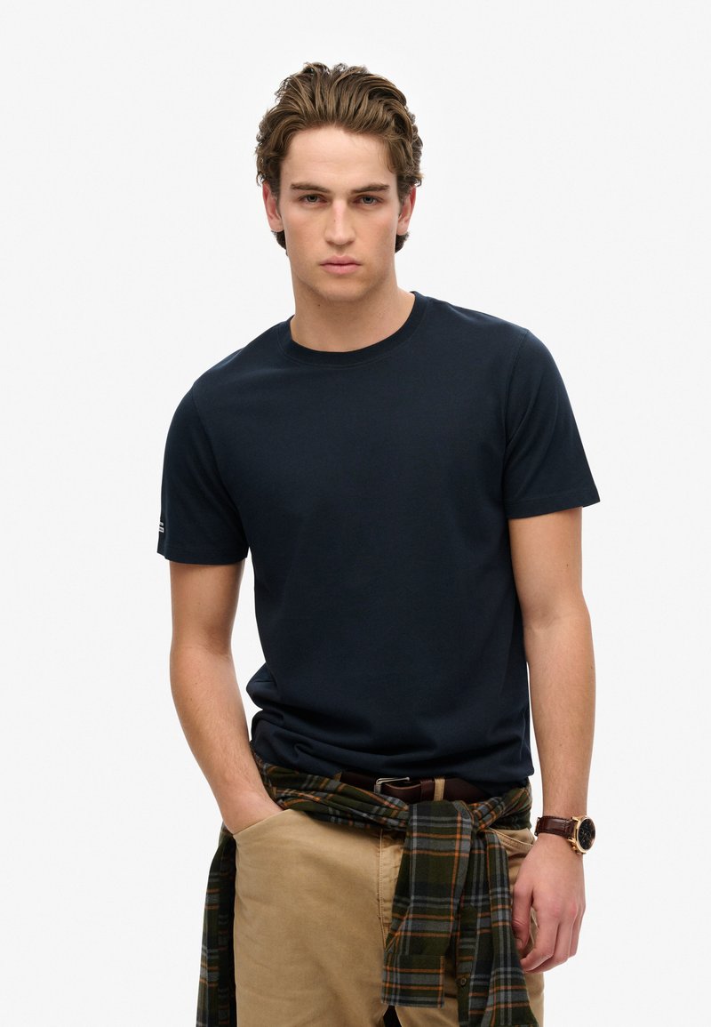 Marineblå crew-neck T-shirt i bomuld med korte ærmer, parret med khakifarvede bukser og en grønternet flannelbundet om taljen. Brun urskov synlig.