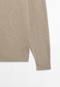 Beige knitted sweater sleeve and lower torso section with ribbed cuffs and hem on a white background.
