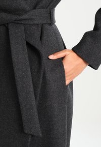 Mano inserita nella tasca di un cappotto di lana grigio scuro con cintura in tessuto abbinata annodata in vita.