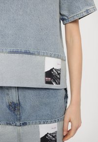 Lichtblauwe denim outfit met twee rechthoekige patches die zwart-wit bergen tonen en de tekst "Moschino Jeans" dichtbij de zijkant van een persoon.