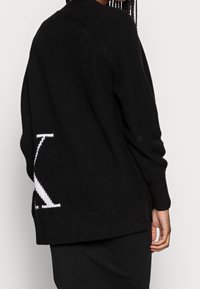 Černý pletený svetr s ribovanou texturou, kulatým výstřihem a bílým logem na zadní straně, s postranními rozparky pro snadný pohyb.