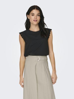 Donna con capelli scuri che indossa un top nero senza maniche con bordo in pizzo e pantaloni beige a vita alta con cintura e fibbia in metallo.