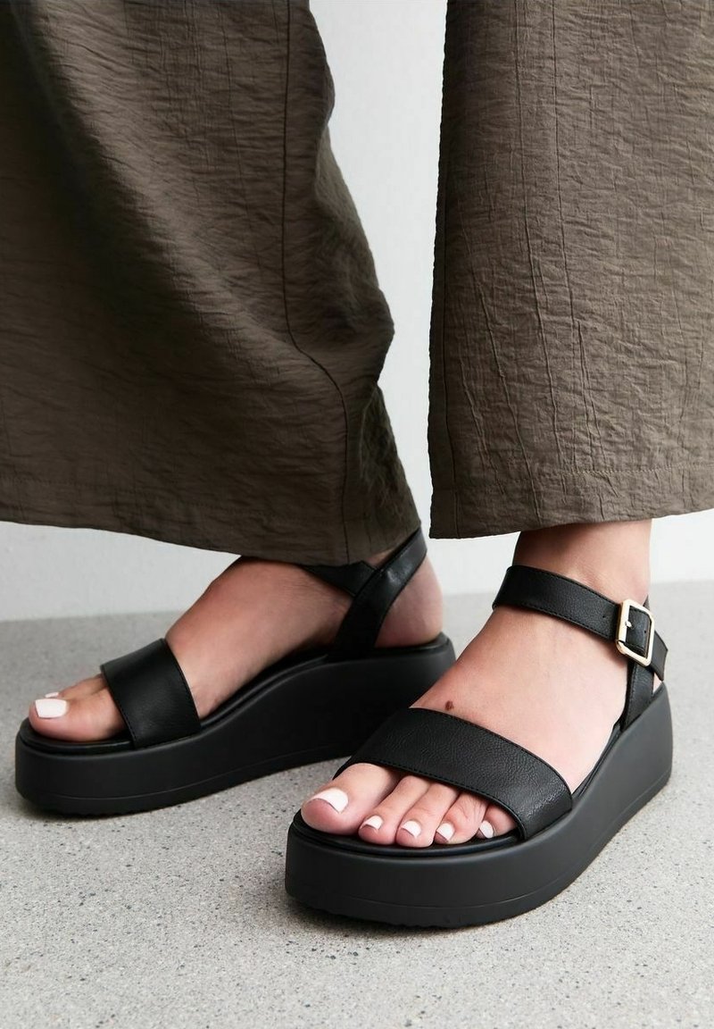 Fötter i svarta platåsandaler med ankelremmar och vitt nagellack, stående på grått golv, klippt nedanför vadens bruna byxor.