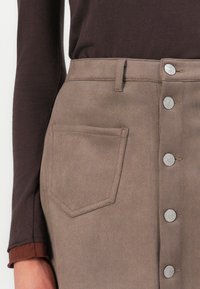 Bruine suède rok met een voorknopsluiting en een opgestikte zak, met zilverkleurige details en een gladde textuur.