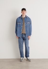 Világoskék farmerdzseki, két mellzsebbel, egy bézs ing felett viselve. A szettet slim-fit kék farmer és fekete-fehér sportcipő egészíti ki.