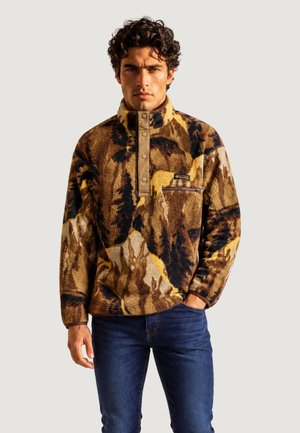 Uomo con capelli ricci che indossa un pullover in pile marrone e giallo a motivo e jeans blu, in piedi con una mano in tasca.