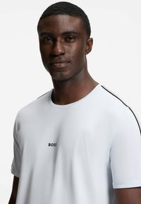 Valkoinen lyhythihainen T-paita, jossa on pyöreä kaula-aukko, ja jossa on musta "BOSS" -logo rinnassa sekä kontrastoivat raidat hartioilla.