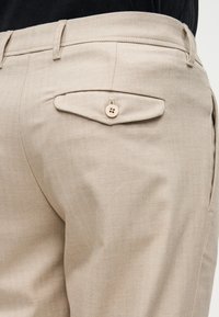 Pantalon beige à motif chevron avec une poche arrière unique fermée par un bouton. Le tissu est lisse et la coupe est ajustée.