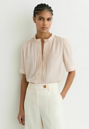 Žena s vrkočmi, ktorá má na sebe svetloružovú textúrovanú blúzku s krátkym rukávom a biele vysoký pás nohavice s jedným gombíkom.