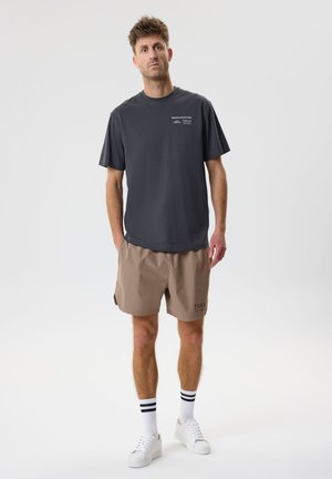 Mees kannab tumehallil üle suure T-särgi ja beežid lühikesed püksid, seisab käed taskutes ühetaustalise hele tausta ees.