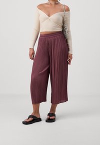 Béžový top bez ramínek s ribkováním, zkombinovaný s burgundskými plisovanými culotami. Černé ploché sandály dotvářejí outfit. Viditelná stříbrná kabelka přes rameno.